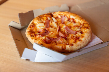 A pizza with ham and pineapple sits in a cardboard box on a table