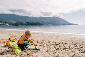 Little girl squats on the seashore and digs the sand with a shovel holding a plastic mold in her...