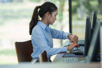 Young businesswoman sitting at her desk in a bright office, typing on a computer keyboard while checking the time on her smartwatch, managing her workload efficiently
