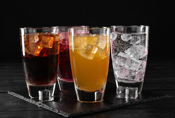 Refreshing soda water of different flavors with ice cubes in glasses on black wooden table