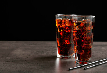 Cold cola with ice cubes in glasses and drinking straws on grey table against black background. Space for text