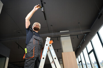 Man install a spot track LED light in the ceiling at the new office. Electrician staying on ladder....