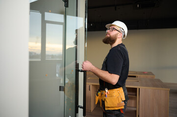 Bearded worker installing glass door in the new office. Construction worker installing door in a new house construction site. Builder in uniform adjusting glass door in new apartment.