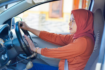 An asian muslim woman in a red hijab and orange outfit is driving a car, looking focused while holding the steering wheel.