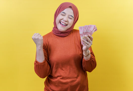 A cheerful asina muslim woman in a hijab celebrates while holding Indonesian rupiah, expressing happiness and success.