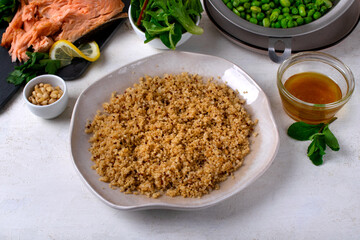 Cooked quinoa on a ceramic plate on a white table. Around it are other ingredients for making a salad: smoked trout, edamame, green peas, dressing, pine nuts, mint and lettuce leaves