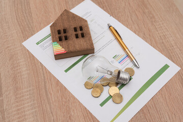 Wooden house model, light bulb and coins on energy efficiency documents, home energy savings and...