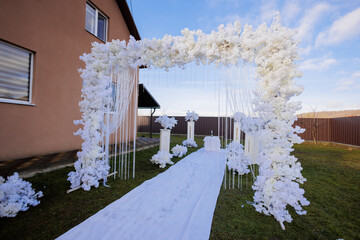 A white archway with white flowers and white flowers in the foreground. The archway is in front of a house