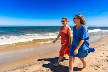 Summer vacations. Two beautiful middle aged women walking on sandy beach enjoying sunny and windy...