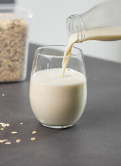 Close up of a female hand pouring oat milk into a glass on grey kitchen table. Vegan and non-dairy alternative milk
