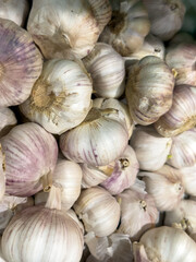 Close-up of fresh garlic bulbs with white and purple skins piled together. The bulbs vary in size, with some having dry stems that show natural textures and details.