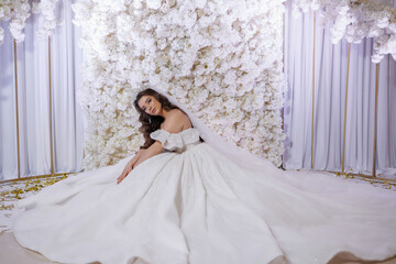Fototapeta premium A woman in a long white dress is sitting on the floor in front of a wall of white flowers