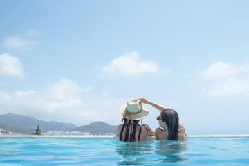 Two beautiful young women are inside a blue and crystalline water pool. They have long, brown and wet hair and one of them puts hat to the other. Fun and summer holiday concept
