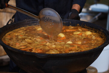 The cook stirs a thick, delicious soup in a large cast iron cauldron with a slotted spoon. National Asian food. Cooking meat dishes outdoors. Selective focus.