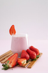 Vertical image of strawberry smoothie in a glass cup next to scattered strawberries on a light...