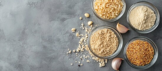 Dried garlic flakes granules and powder in clear glass bowls arranged on a grey textured surface with raw garlic cloves scattered around