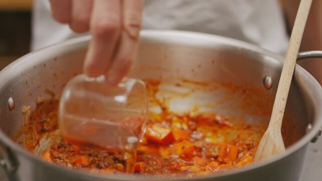 Koch gie&szlig;t Br&uuml;hwasser aus Glas mit Hand im Close Up in Aluminium Topf in ein Hackfleisch, Karotten, Kartoffel Gemisch