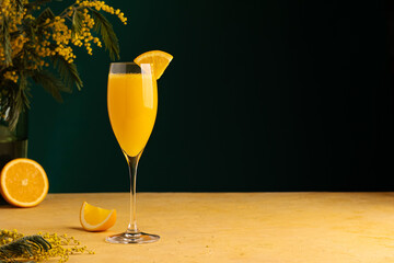 Mimosa cocktail in a champagne glass garnished with a triangle piece of orange. Alcoholic drink made from freshly squeezed orange juice and sparkling wine. Yellow table, dark background. Copy space 