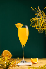 Mimosa cocktail in a champagne glass garnished with a piece of orange. Alcoholic drink made from fresh orange juice and sparkling wine. Yellow table, dark green background. Copy space 