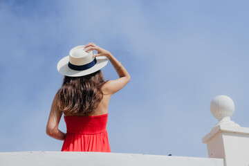 A young teenage woman is on her back with the blue sky in the background. She is holding an elegant white hat and she wears a red dress. The summer wind moves her dark and long hair