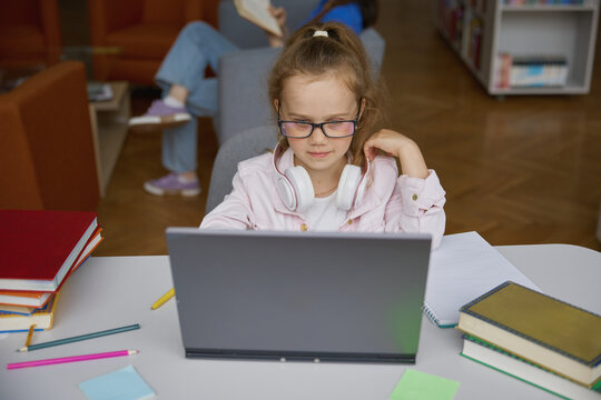 Happy little girl studying via internet using laptop in library - Powered by Adobe