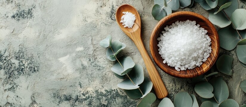 Wooden bowl filled with coarse salt beside a wooden spoon on a textured gray background with green eucalyptus leaves arranged artistically - Powered by Adobe