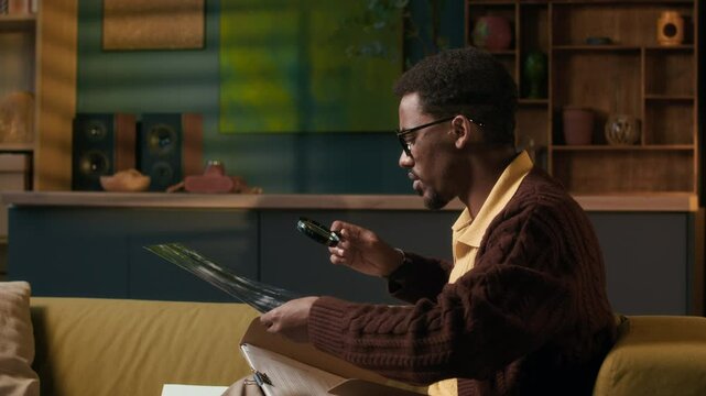 Handheld shot of young male specialist of African American ethnicity analyzing pictures under magnifying glass at home studio