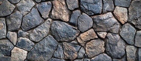 Fototapeta premium Textured dark stone wall with irregularly shaped gray and brown stones creating a natural rustic background emphasizing depth and variation.