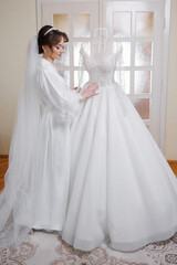 Fototapeta premium A bride is standing in front of a wedding dress, which is displayed on a mannequin. The bride is wearing a white gown and a veil. Concept of anticipation and excitement for the upcoming wedding