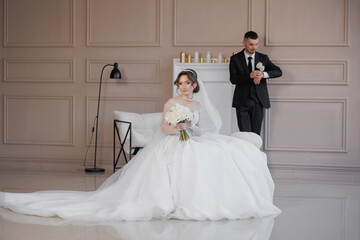 Naklejka premium A bride and groom are posing for a picture in a room with white walls. The bride is wearing a white dress and holding a bouquet of flowers. The groom is wearing a suit and tie