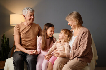 Grandparents and two young children sit closely on a couch, sharing smiles and laughter in a warm,...