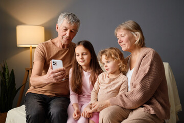 Grandparents and two young girls sit closely on a couch, sharing laughter and smiles while engaging...