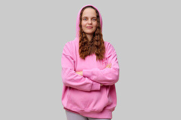 Obraz premium A woman with long, wavy hair wears a pink hoodie and stands confidently with arms crossed. Her expression is relaxed and approachable, set against a simple neutral backdrop