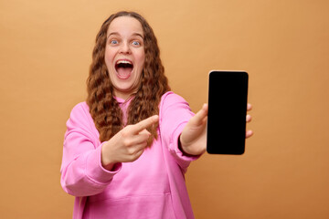 A young woman with wavy hair, wearing a pink hoodie, enthusiastically points at her smartphone,...
