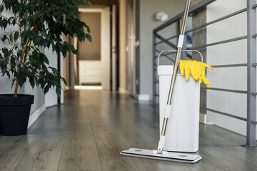 Cleaning supplies: mop, gloves, bucket with wringer sitting on wooden laminate floor in modern living room, wet equipment tools, housekeeping concept. Emphasizing home sanitation, cleanliness, hygiene