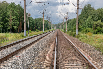 Naklejka premium Straight railway tracks cutting through the forest and going off into the distance under a cloudy sky. Overhead power lines create a sense of symmetry and travel through nature.