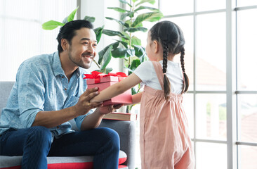 Obraz premium Asian cute little daughter giving gift box, making surprise for dad, greeting young man with father's day or birthday. Selective focus on father smiling and sitting on sofa at home
