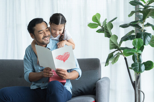 Little daughter kid making surprise present for her father for father's day or birthday, giving dad a handmade card with heart and reading together on sofa at home. Asian family celebrating holiday