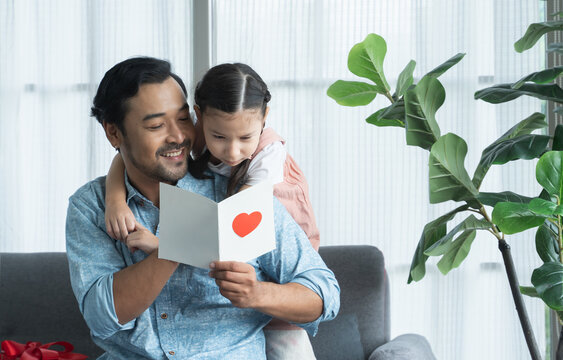 Little daughter kid making surprise present for her father for father's day or birthday, giving dad a handmade card with heart and reading together on sofa at home. Asian family celebrating holiday - Powered by Adobe