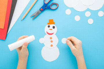 Little child boy hands creating and gluing snowman shape from white paper on light blue table...