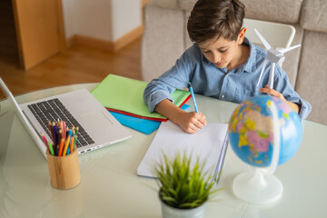 Schoolboy taking notes in his notebook while studying renewable energy and sustainability with a small wind turbine and a globe on his desk at home