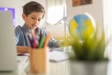 Young schoolboy drawing and learning about renewable energy while interacting with a model wind turbine and globe at his desk, surrounded by colored pencils and a laptop