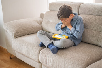 Frustrated child sitting on the couch covering his face with his hands, crying inconsolably after...