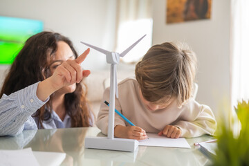 Mother pointing at a small wind turbine model while explaining renewable energy concepts to her son, who is diligently taking notes, fostering stem education and environmental awareness at home © Nanci