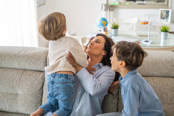 Obraz premium Mother is holding her youngest son, playing with him on the sofa, while her older son is sitting next to them, enjoying the family moment in their living room