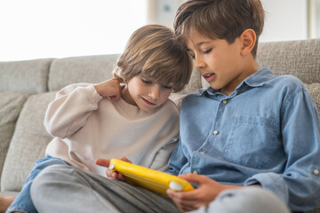 Two young brothers are sitting on a comfortable sofa, engrossed in a video game on a handheld console, enjoying their time together in the living room