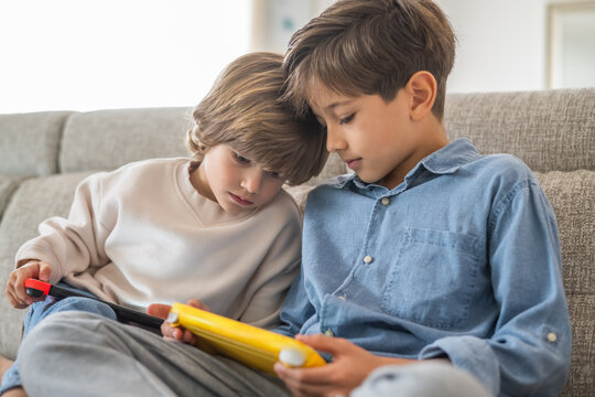 Two young brothers are sitting close together on a comfortable sofa, engrossed in playing a video game on a handheld console, demonstrating sibling bonding and shared enjoyment of technology