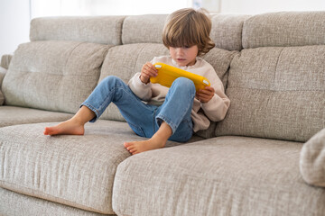 Happy child sitting barefoot on a comfortable sofa at home playing videogames on a yellow portable...