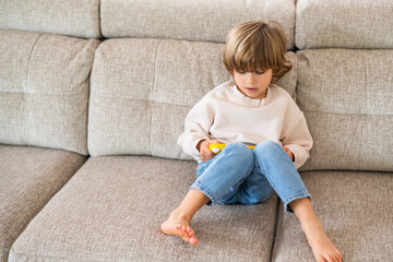Barefoot child sitting comfortably on a sofa, engrossed in playing with a handheld video game...