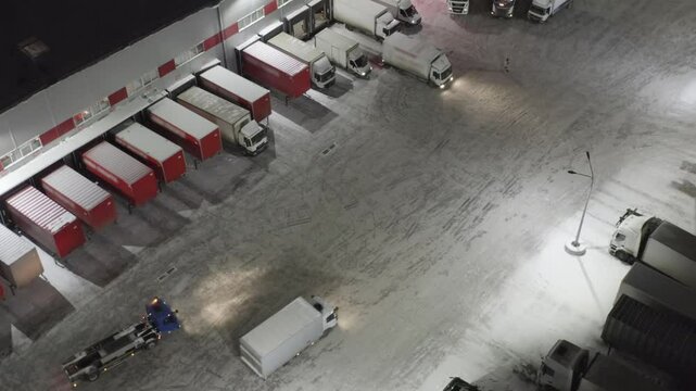 Aerial View of Logistics Center with Trucks Loading at Night. Winter Snowy Scene of Warehouse Distribution Hub with Cargo Containers and Moving Semi Trailers.